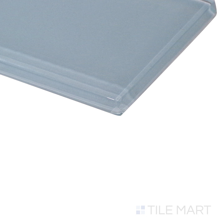 A smooth pastel blue glass tile catches the light from an angled perspective, emphasizing its glossy finish and soft reflections.