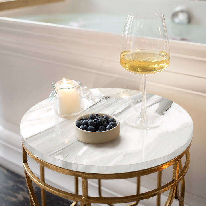 Round marble table with a glass of white wine, bowl of blueberries, and lit candle on a bathtub.
