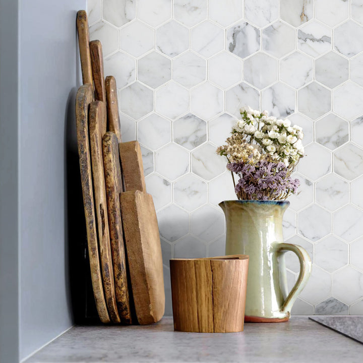 Wooden cutting boards and a ceramic pitcher with flowers on a kitchen counter against a marble hexagonal tile backsplash.