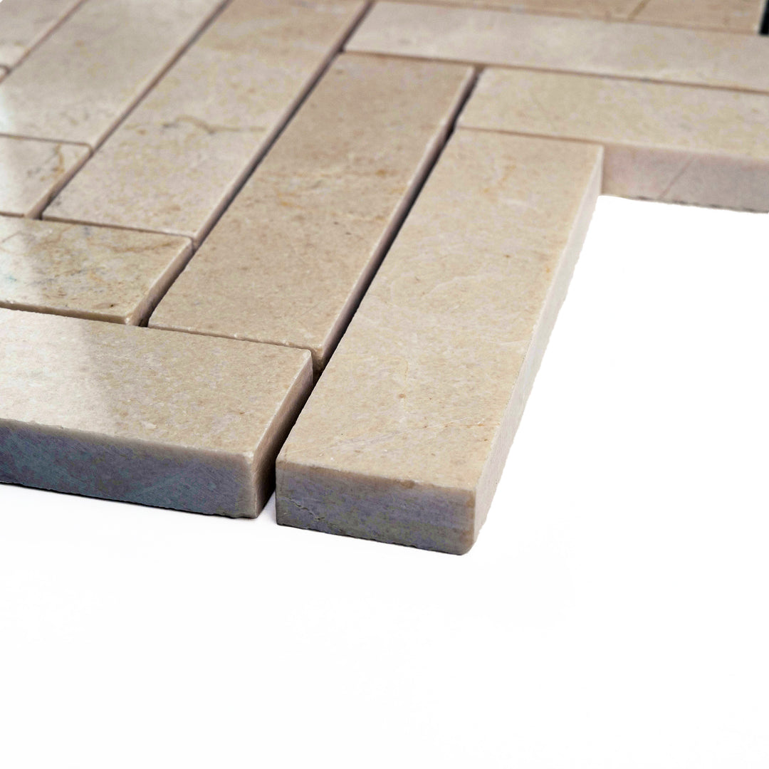Polished 1x4 herringbone mosaic in Crema Marfil marble, corner-shot to reveal its high-sheen finish and elongated V-pattern detail.