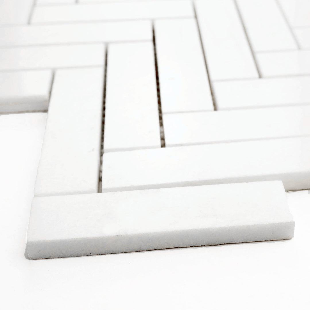 Polished 1x4 herringbone mosaic in Thassos White marble, corner view accentuates reflective finish and graceful pattern layout.