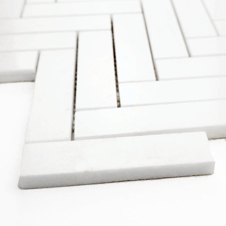 Polished 1x4 herringbone mosaic in Thassos White marble, corner view accentuates reflective finish and graceful pattern layout.